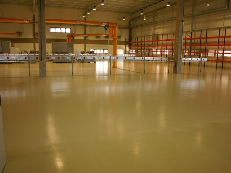 Galpão amplo com piso epóxi bege liso, iluminação industrial e estruturas metálicas de estocagem ao fundo.