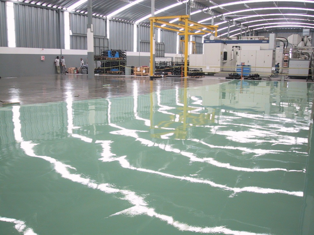 Área industrial ampla com piso epóxi verde refletivo, mostrando máquinas, estruturas metálicas e iluminação no teto.