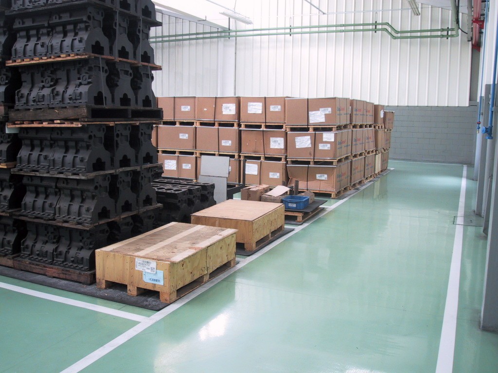 Galpão industrial com piso epóxi verde claro brilhante, contendo pallets de caixas e peças metálicas empilhadas.