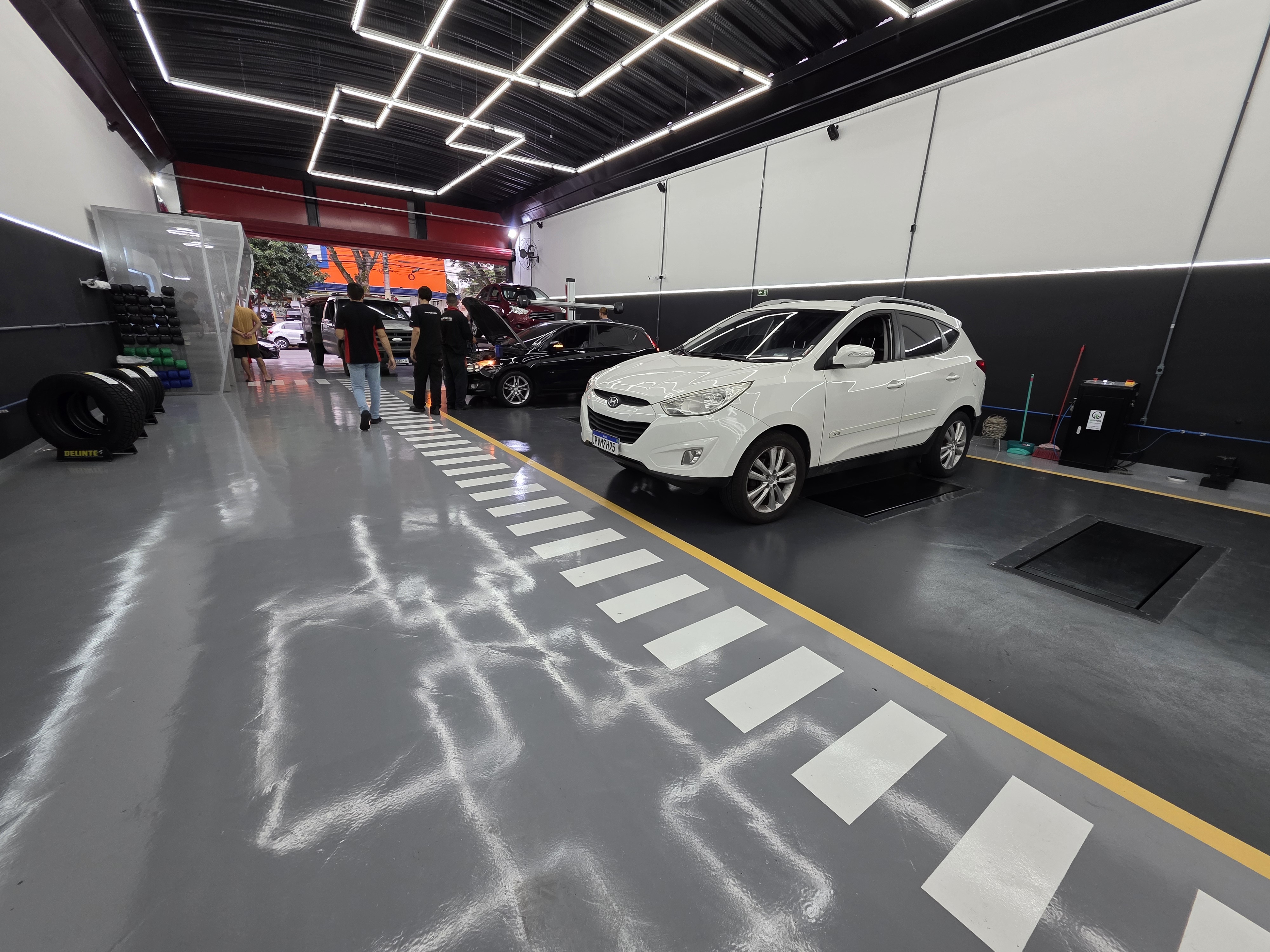 Área de serviço de oficina com piso epóxi cinza, equipamentos de alinhamento e balanceamento distribuídos pelo espaço.