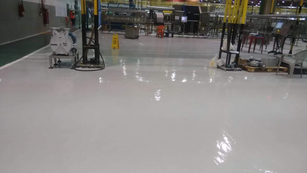 Piso industrial com acabamento brilhante recém-executado, localizado em área fabril com equipamentos e sinalização de segurança.