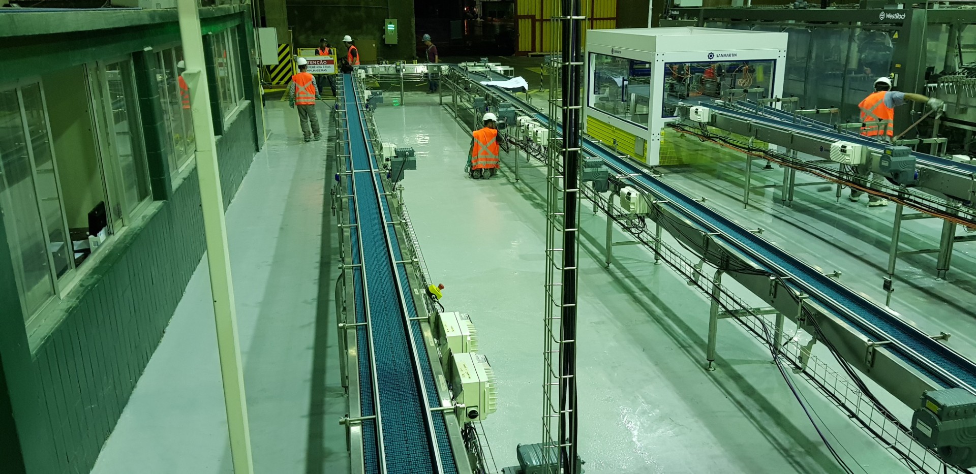 Equipe aplicando revestimento no piso de uma linha de produção industrial, com máquinas e esteiras ao redor.