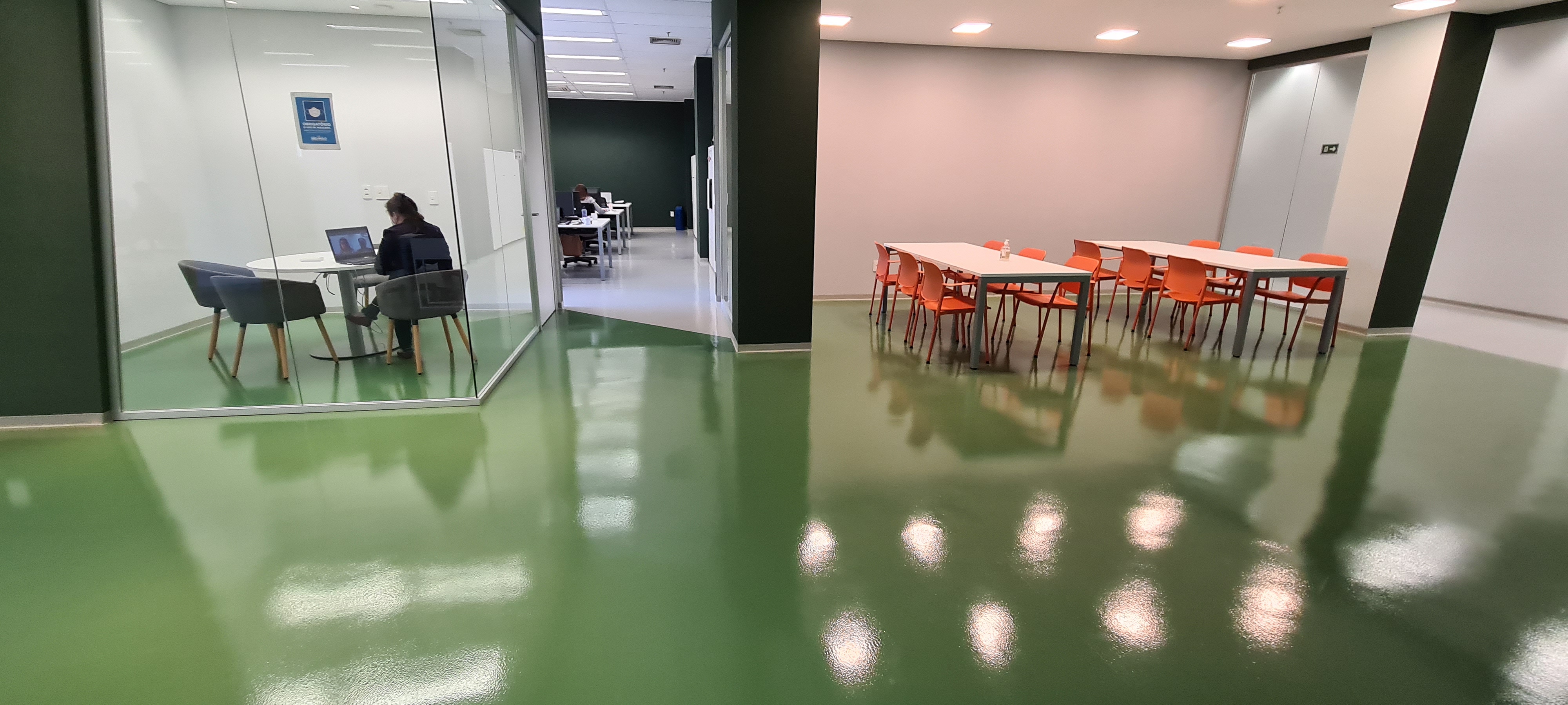 Escritório corporativo com piso epóxi verde brilhante, sala de reunião envidraçada e área de convivência com mesas e cadeiras laranjas.
