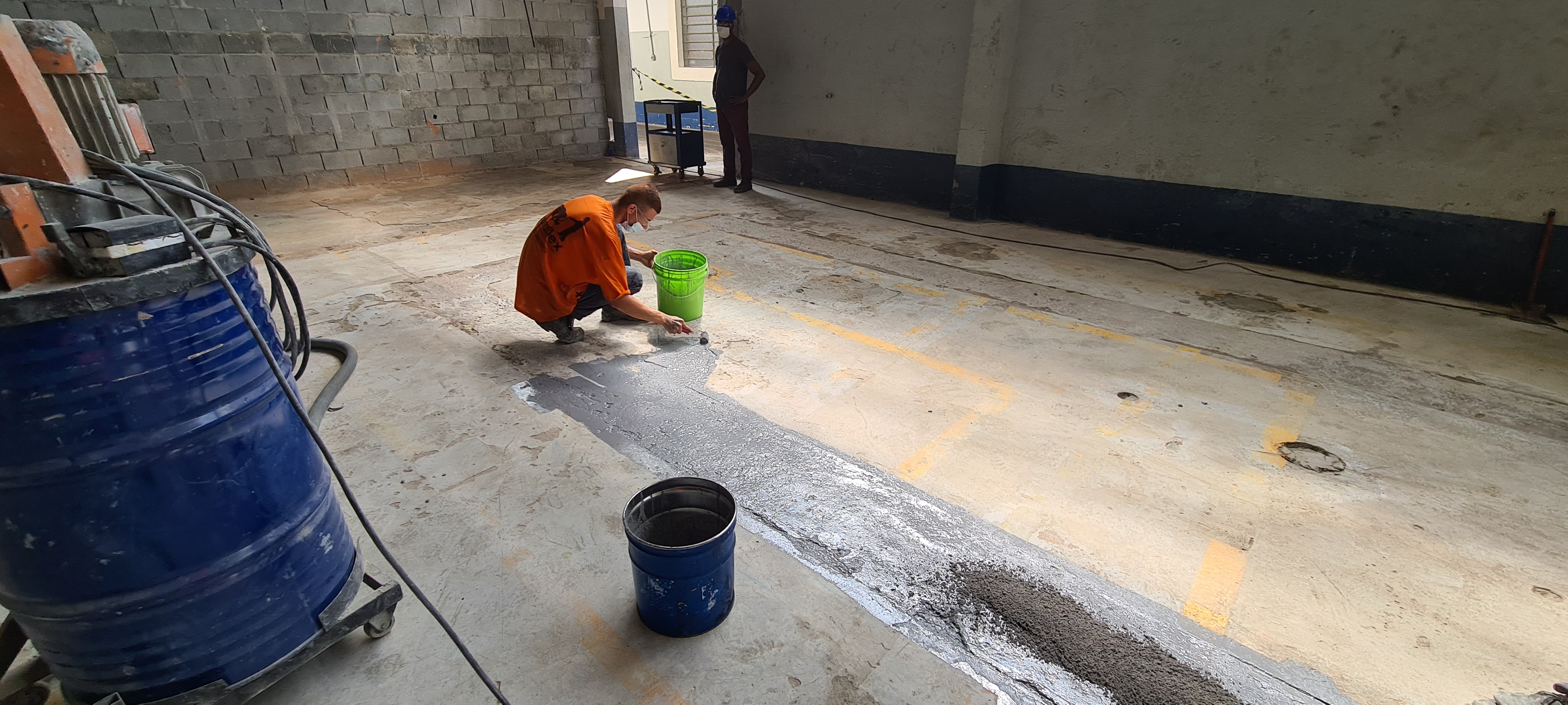 Reparo de piso industrial com argamassa epóxi já nivelada, faixa de demarcação amarela visível ao fundo.