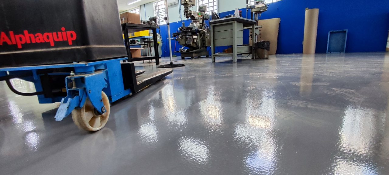 Piso industrial cinza brilhante em setor de montagem, com paleteira e bancadas azuis.