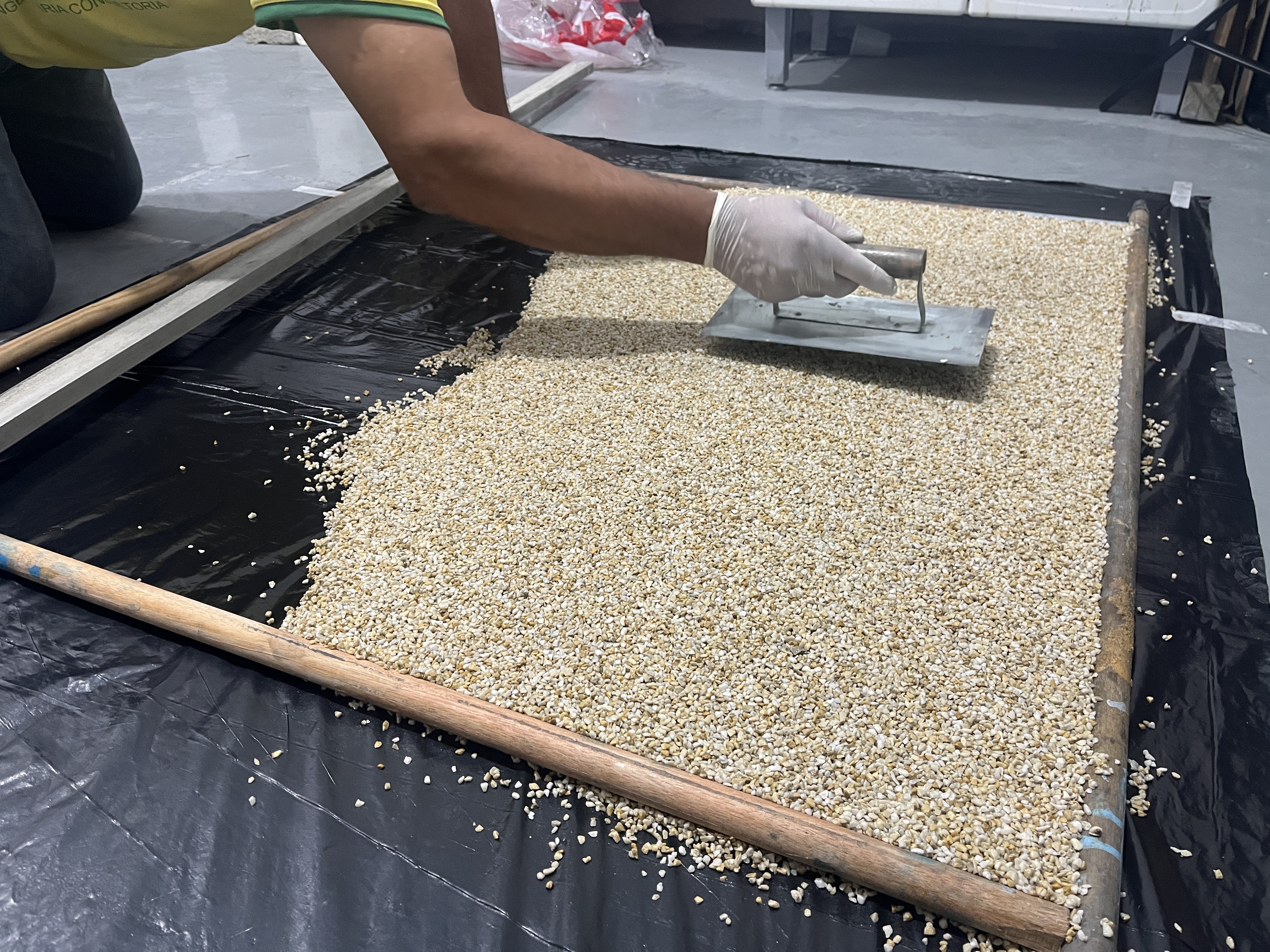 Aplicador espalhando grânulos de quartzo bege sobre resina em painel de fôrma, preparando trecho de piso drenante.