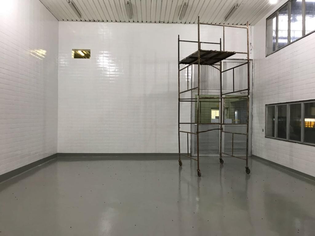 Salão industrial vazio com paredes brancas revestidas de azulejo e piso epóxi cinza recém-instalado.