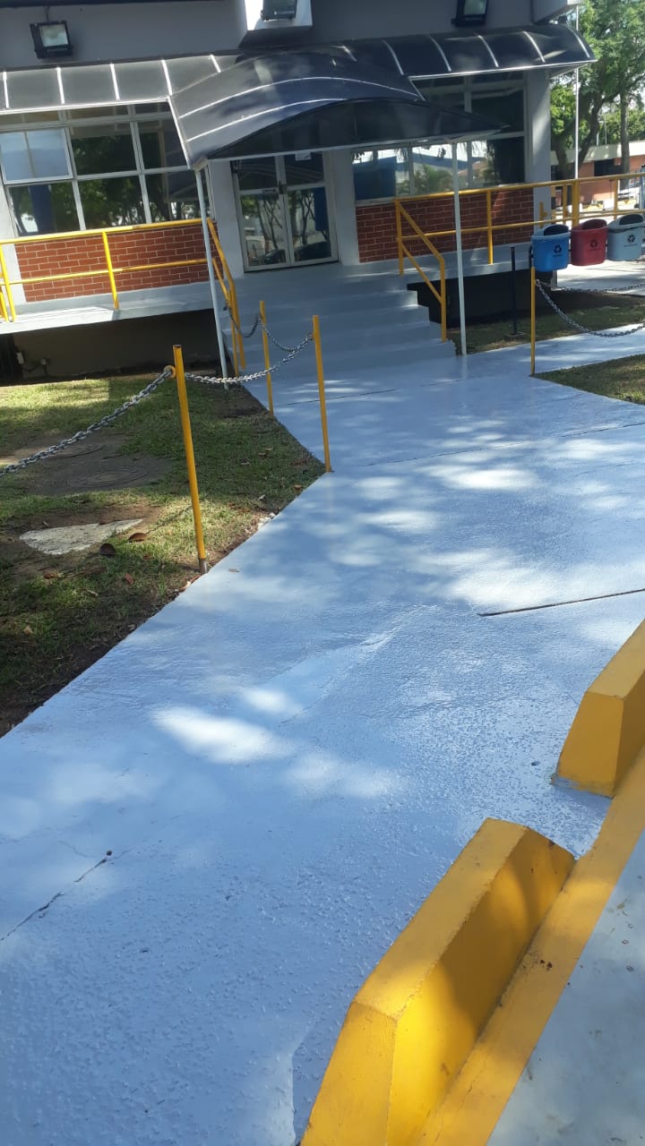 Passarela de acesso com piso em uretano azul-claro, corrimãos amarelos e coletores de reciclagem coloridos ao fundo.