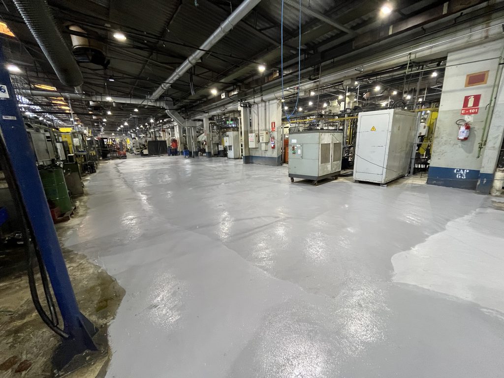 Corredor de fábrica com piso resinado cinza claro recém-aplicado entre máquinas e painéis elétricos.