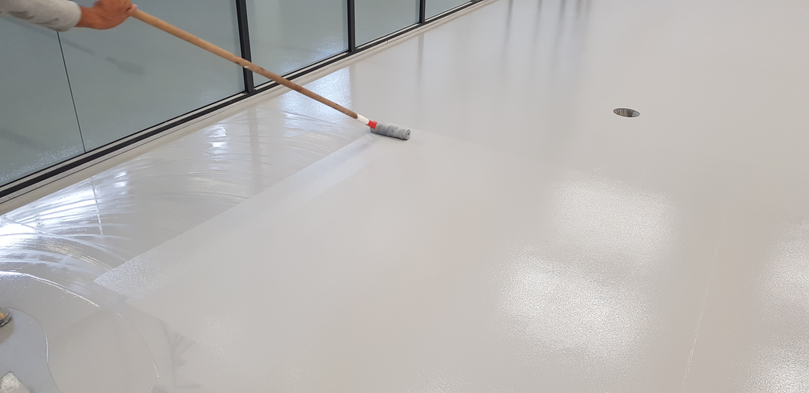 Aplicação de tinta epóxi branca com rodo sobre piso de ambiente envidraçado.