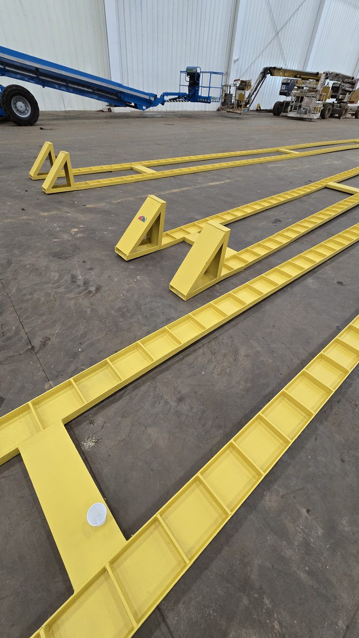 Peças metálicas com pintura anticorrosiva amarela alinhadas no piso de um galpão industrial, com plataforma elevatória ao fundo.